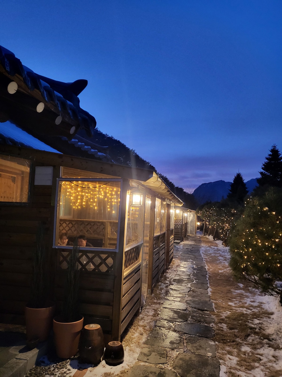 A gentle pathway lined with stone connects wooden structures, illuminated by warm string lights. The twilight sky casts a soft glow, highlighting the serene surroundings. Decorative plants are placed near the entrance, creating an inviting atmosphere as trees stand quietly in the background.