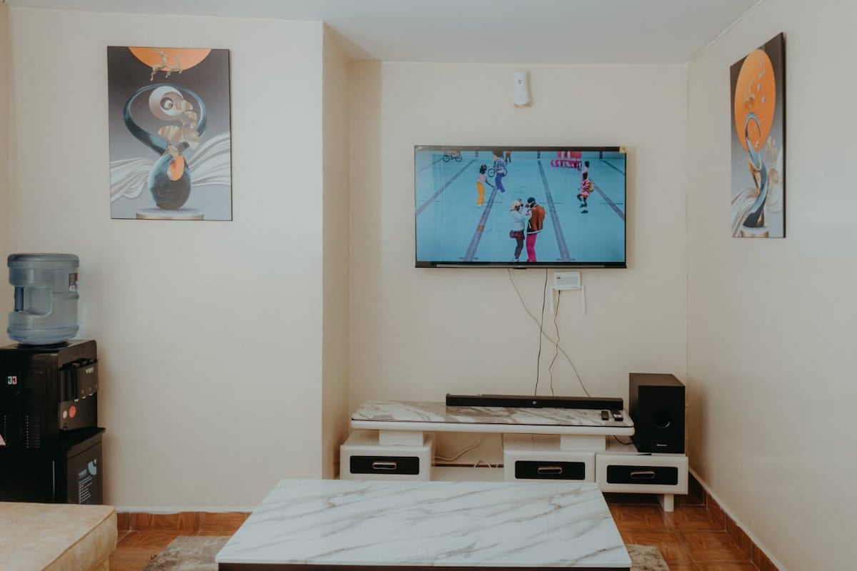 A living area is presented with light-colored walls and wooden flooring. A flat-screen TV is mounted on the wall, showing a sports program. Below, a low coffee table with a marble top complements a stylish console underneath, flanked by two artworks on adjacent walls.