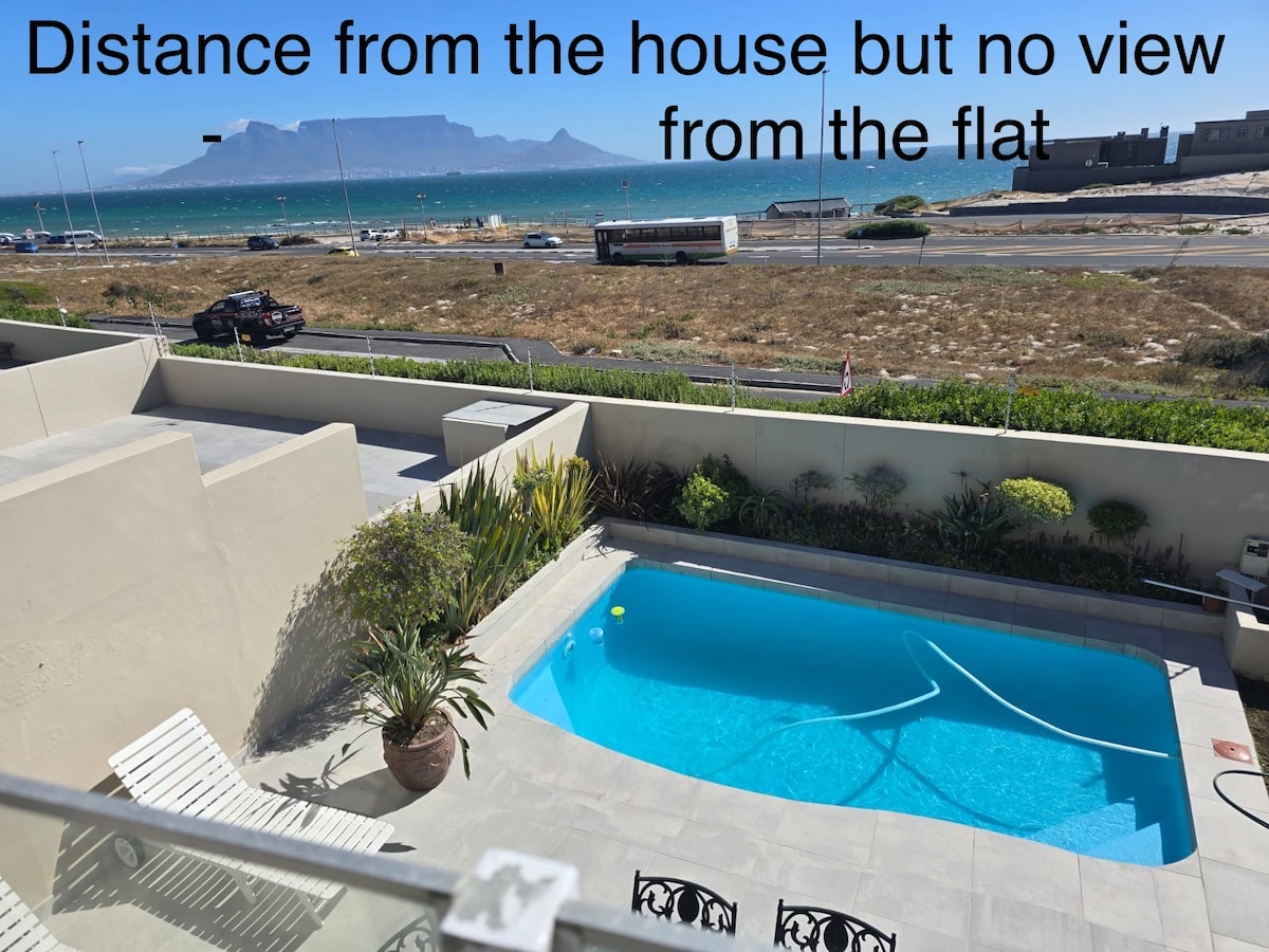 The image captures a clear view of an outdoor area featuring a small rectangular pool set on light grey tiles. Lush greenery surrounds the pool, and lounge chairs are arranged on one side. A distant ocean and mountain backdrop can be seen beyond the property.