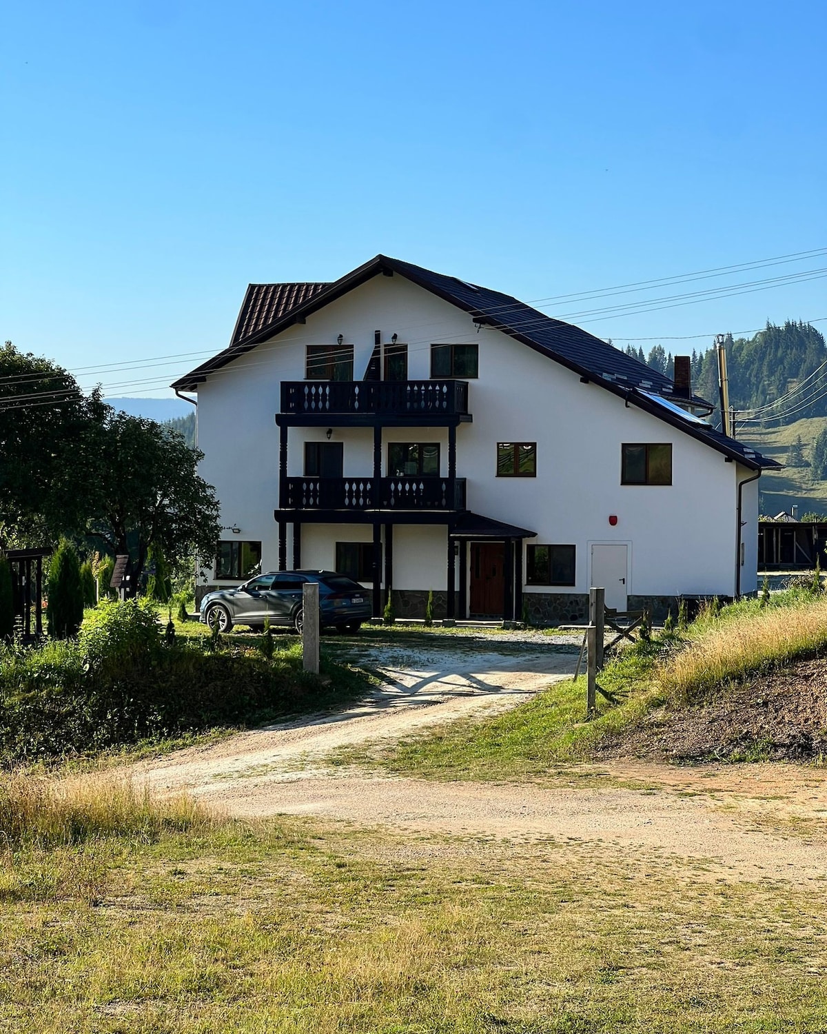 The exterior of a three-story house is visible, featuring a dark roof and multiple balconies. The building is surrounded by greenery and opens onto a gravel pathway leading to a parking area. The serene landscape includes distant trees and mountains under a clear blue sky.