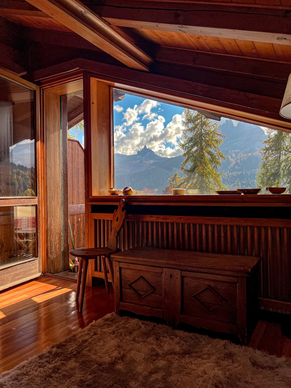 Natural light fills a cozy corner of the chalet, highlighting a wooden chair beside an antique chest. Large windows offer a stunning view of the mountains and sky, framed by warm wooden paneling that adds to the inviting ambiance.