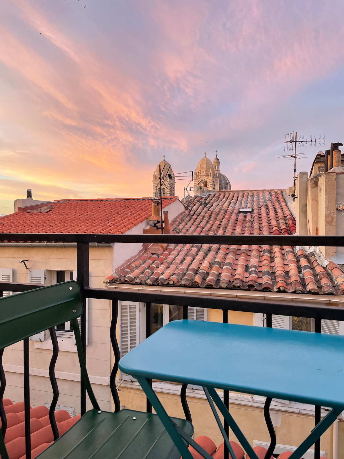 A balcony view showcases terracotta rooftops under a colorful sunset sky. In the foreground, a green metal chair and small blue table are visible, with the domes of a church peeking over the rooftops, creating a serene outdoor space.