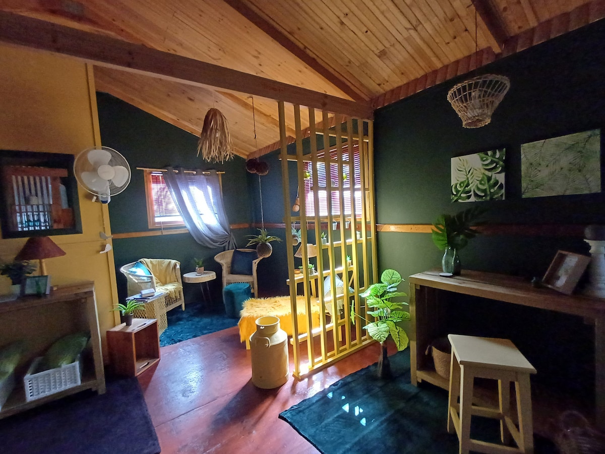 A cozy interior space is highlighted with wooden beams and warm-toned walls. Green plants add a touch of nature, while a weave basket and artwork accent the atmosphere. A divider creates separate areas, showcasing comfortable seating and soft textures that invite relaxation.