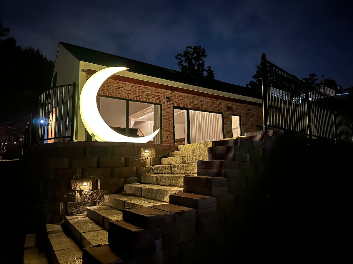 A modern building is illuminated at night by soft lighting, highlighting a distinctive crescent-shaped sculpture near the entrance. The stone steps lead up to the entrance, with large windows reflecting the warm glow from inside the space.