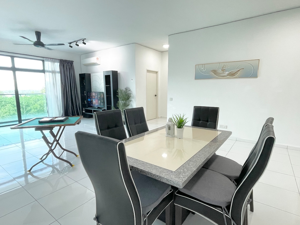 A spacious dining area features a rectangular table surrounded by six upholstered chairs. Natural light streams through large windows, complementing the neutral colour palette. In the background, a living space includes a television and a green view visible through the glass balcony door.