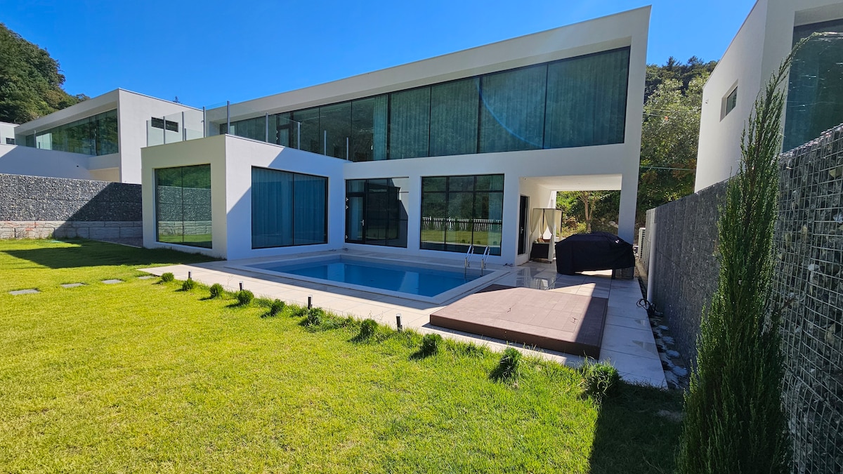 A modern villa is depicted, featuring large glass windows and a sleek design. A private swimming pool is visible in the foreground, surrounded by a well-maintained lawn. A wooden deck is shown adjacent to the pool, with shaded seating areas in the background.