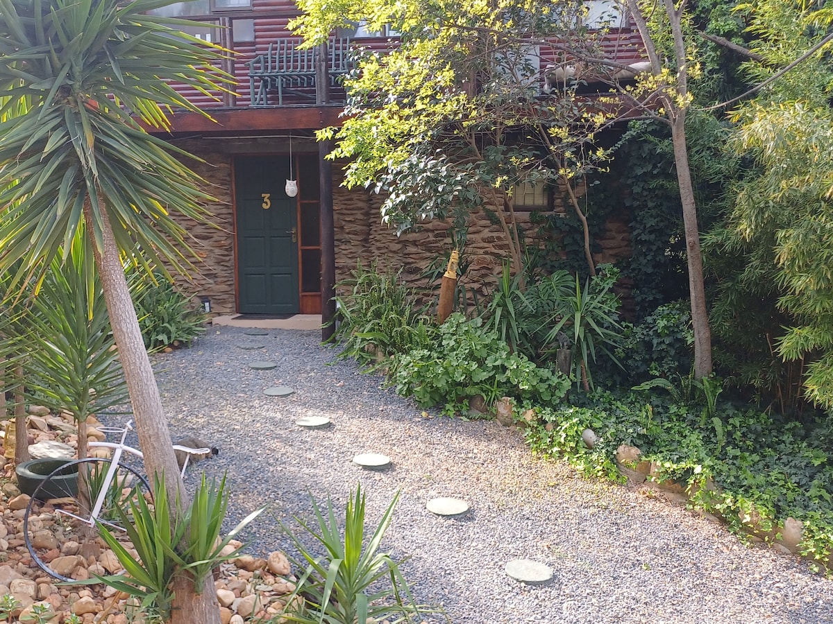 A gravel pathway leads to a stone entrance marked with the number 3. Lush greenery and well-maintained shrubbery surround the entrance, creating a natural ambiance. A small porch with a railing is visible above, adding character to the inviting entryway.
