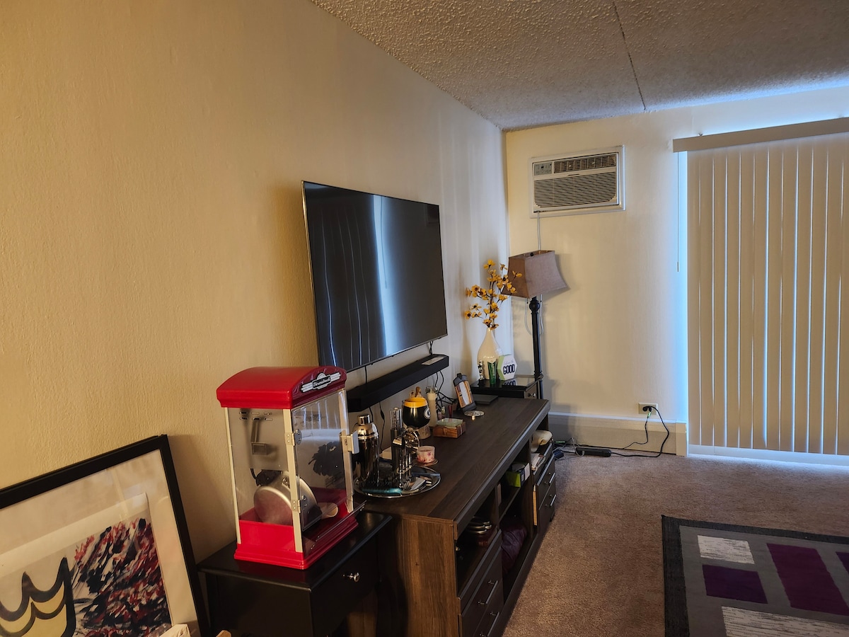 The living area showcases a wall-mounted television above a modern low-profile entertainment unit. A red popcorn machine and decorative items are positioned on the unit. Nearby, a standing lamp provides additional lighting, while vertical blinds allow natural light to filter in.