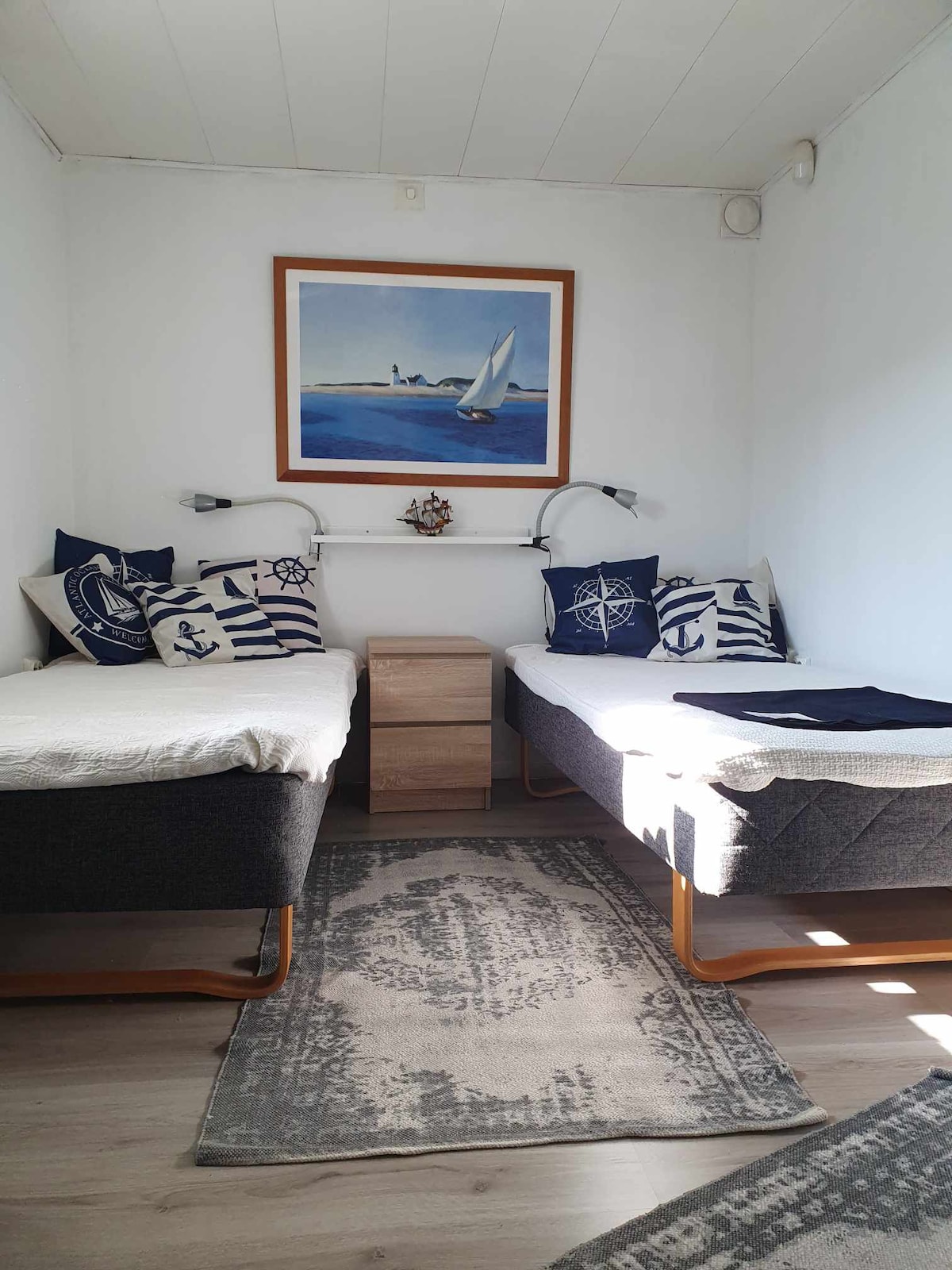 A bright bedroom features two twin beds dressed in nautical-themed linens. Wall-mounted reading lights are positioned above each bed. A wooden nightstand sits between the beds, and a decorative sailing-themed artwork adorns the wall. Natural light filters in through a nearby window.