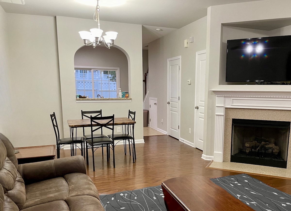 An open living area features a comfortable seating arrangement in front of a gas fireplace. A dining table with four black chairs is positioned nearby. Light walls create a bright space, complemented by a large television mounted above the fireplace.