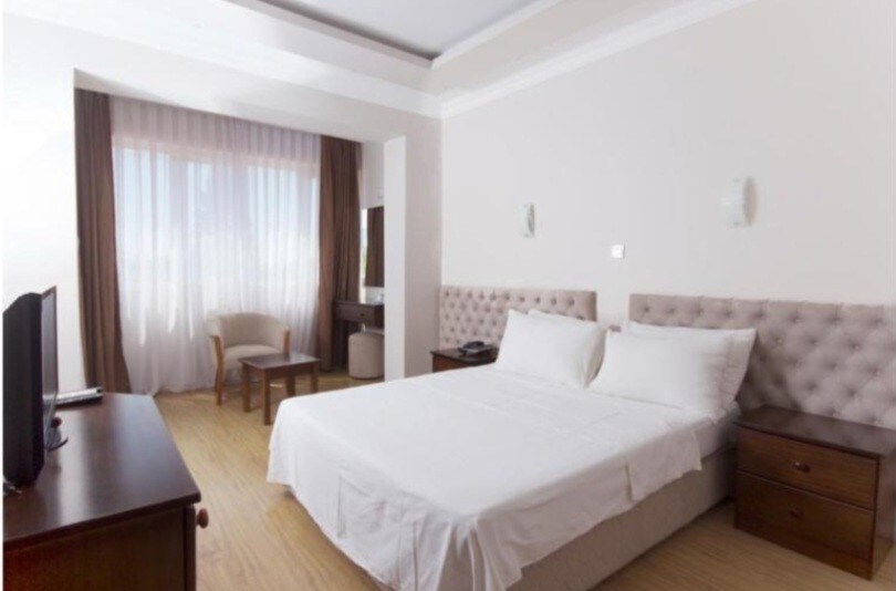 A well-lit bedroom features a spacious bed with a padded headboard and smooth linens. Two bedside tables are positioned on either side of the bed. A chair and a small table are present, and large windows provide natural light, partially covered by sheer curtains.