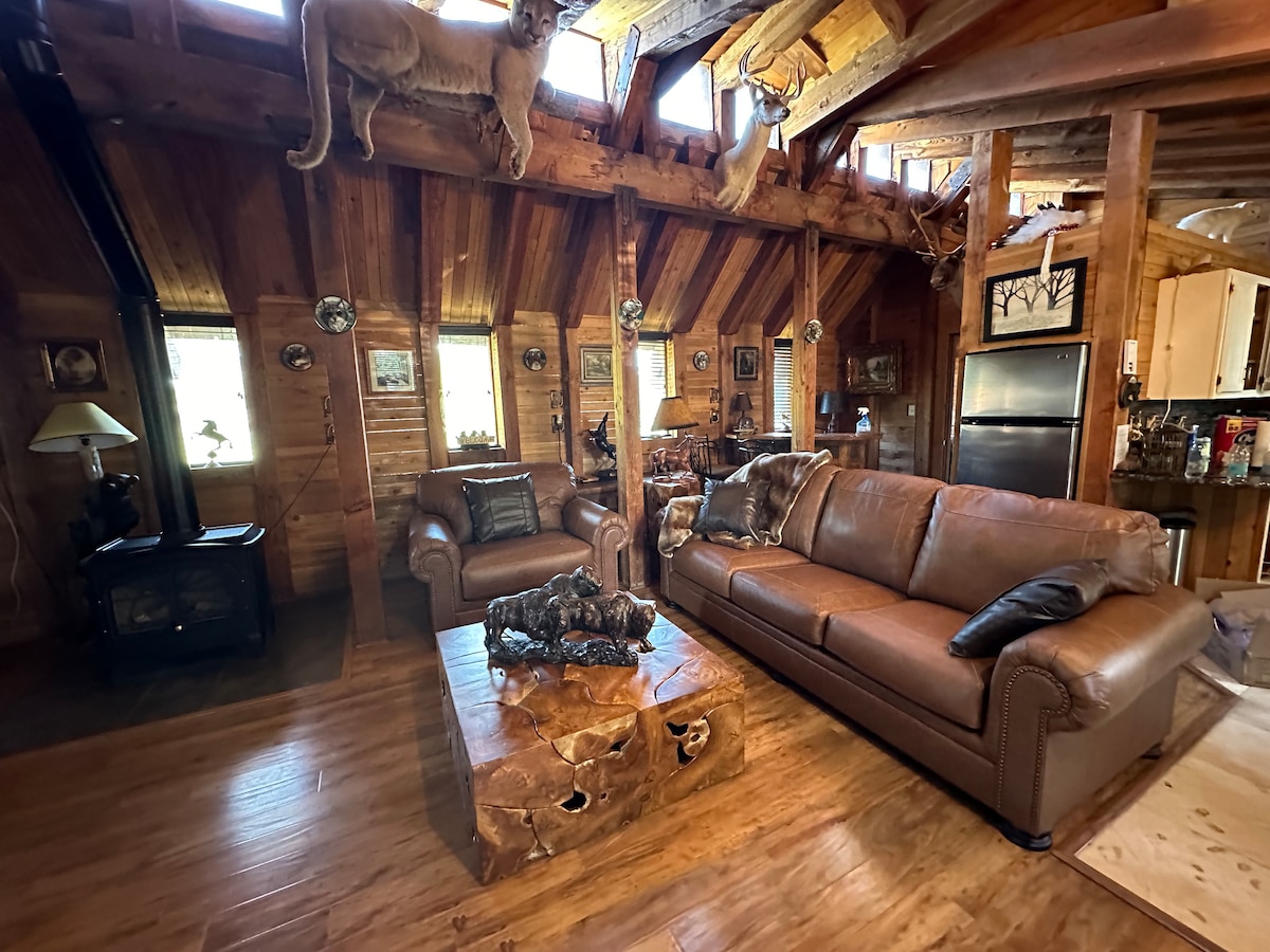 A rustic living area features wooden walls and a high ceiling. A leather sofa and armchair are positioned around a unique wooden coffee table, adorned with animal sculptures. Natural light enters through multiple windows, enhancing the warm ambiance.