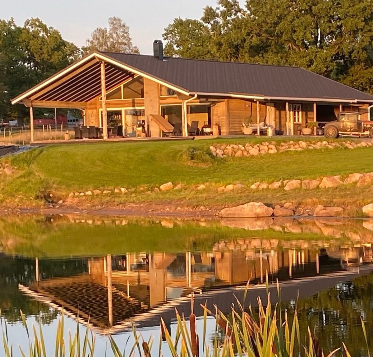 A modern house with a sloped roof is situated by a serene pond, reflecting its structure on the water's surface. Large glass windows provide expansive views of the surrounding greenery. Comfortable outdoor seating is visible on the patio, inviting relaxation in the natural setting.