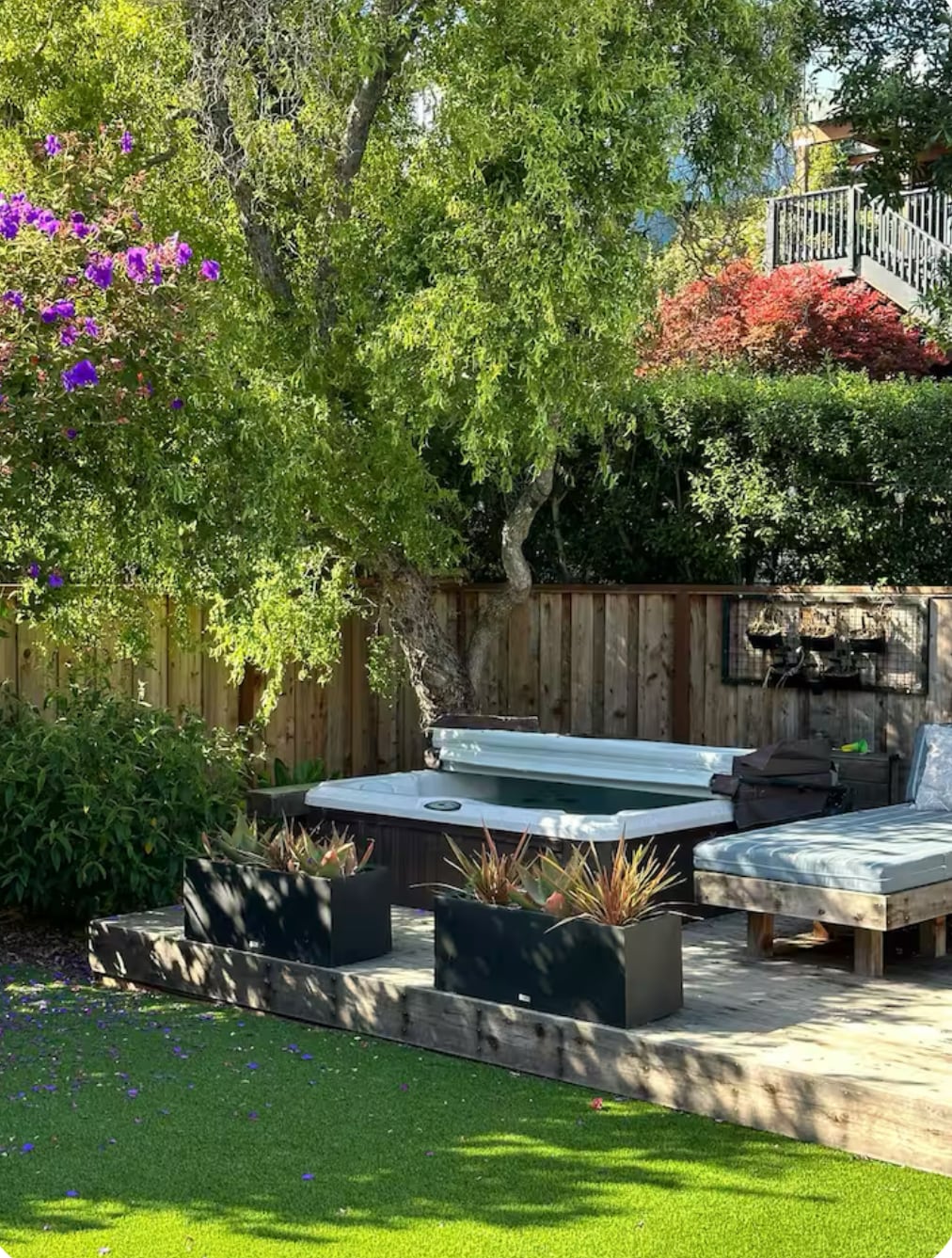 A serene backyard space features lush greenery and flowering plants. A wooden deck is equipped with a comfortable seating area and a gas fire pit, surrounded by planters filled with vibrant foliage. Soft artificial turf enhances the outdoor experience, inviting relaxation.