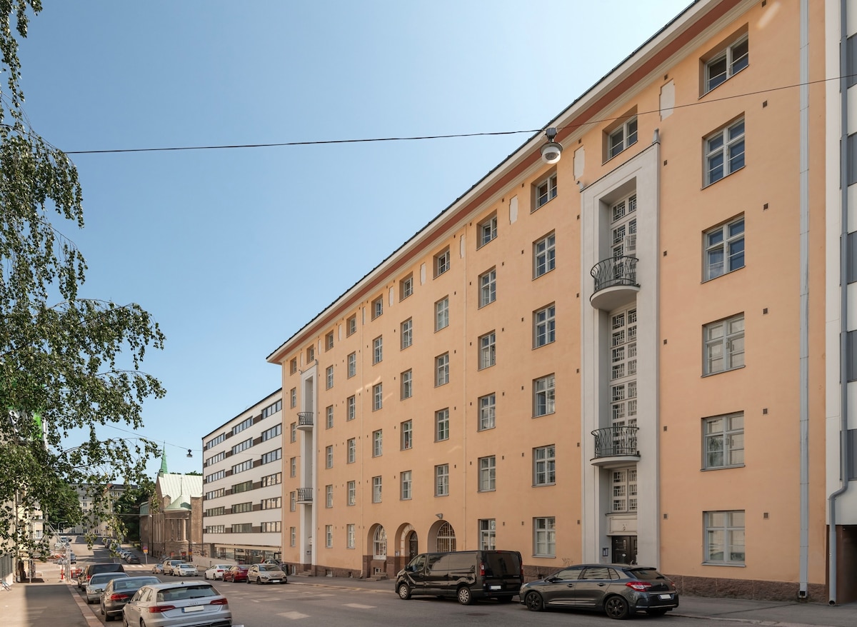 The exterior of a multi-story building is displayed, featuring a light-colored façade with large windows and a modern balcony. The street is lined with parked cars, and a clear blue sky serves as a backdrop. Pedestrians can be seen walking along the sidewalk.