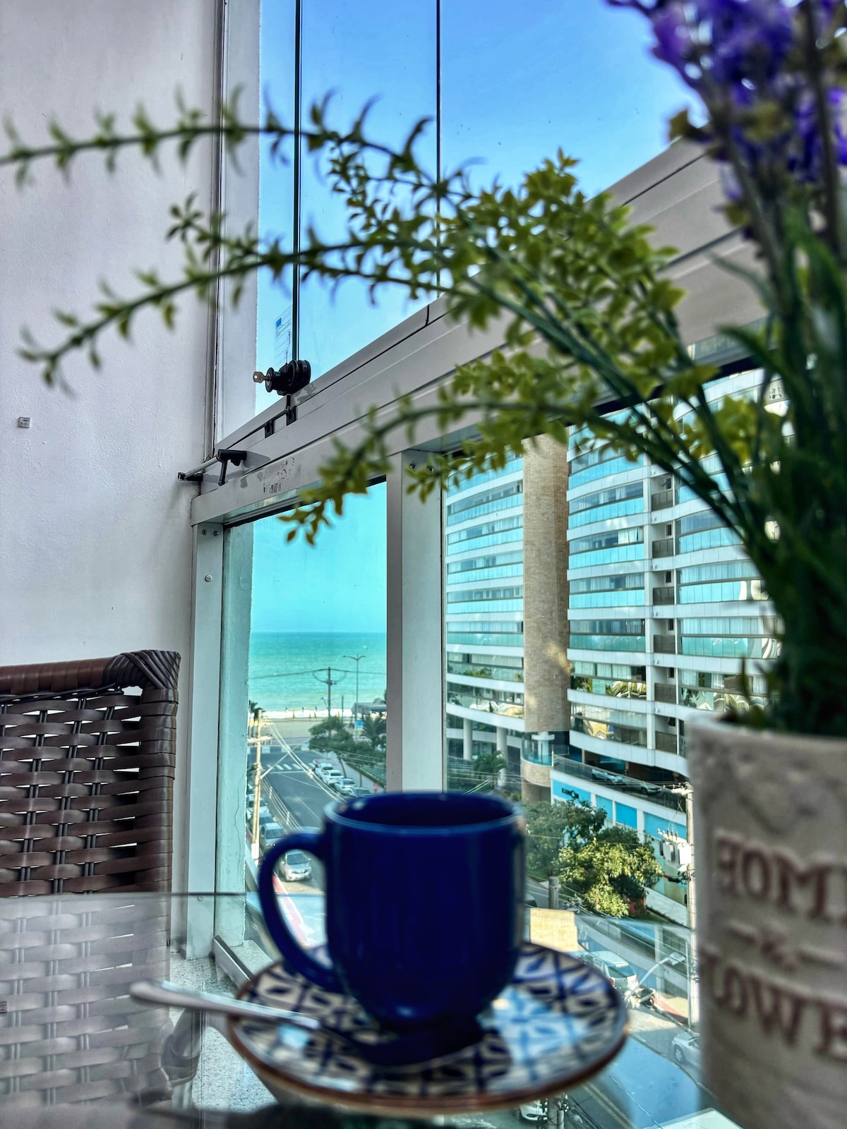 A view from the balcony is captured, featuring large glass windows that open to a coastal scene. A potted plant and a blue cup sit on a glass table, while the ocean and buildings are visible in the background.
