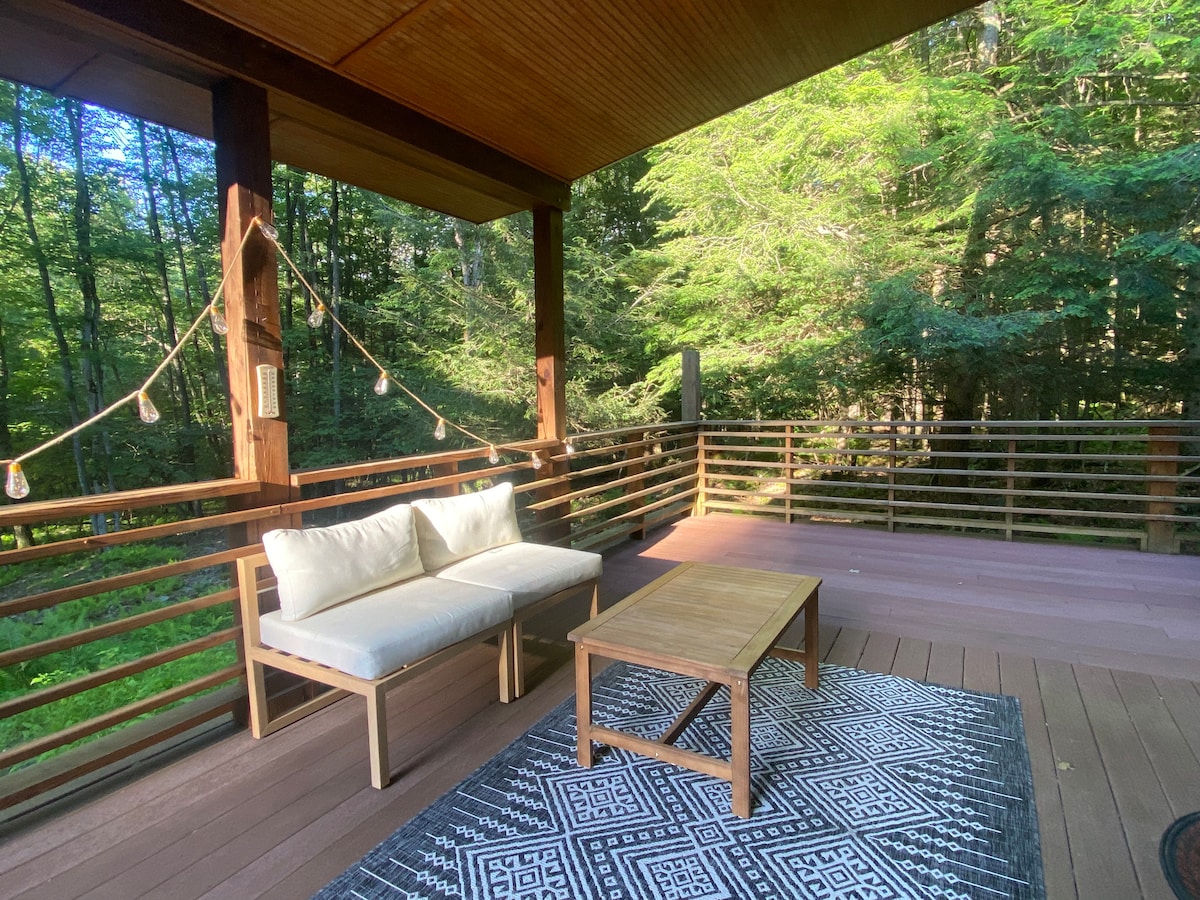 An outdoor deck features a light-colored couch and a wooden coffee table, set atop a geometric-patterned rug. Surrounding greenery and tall trees create a serene backdrop, while soft lighting adds a cozy touch, inviting relaxation in the natural setting.