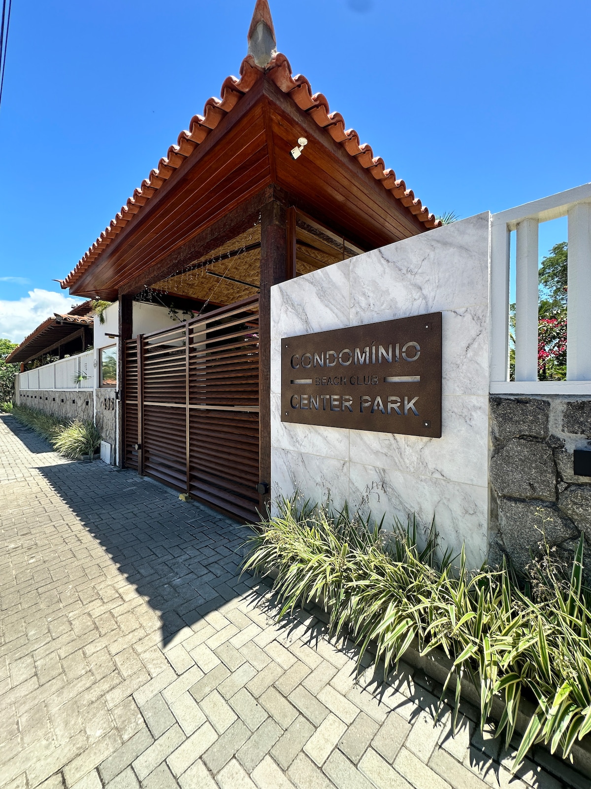 The entrance to the condominium features a contemporary design, showcasing a stone and marble sign that reads 'Condomínio Center Park'. The pathway is lined with neatly arranged greenery, leading up to a wooden gate that blends with the classic roofing style.