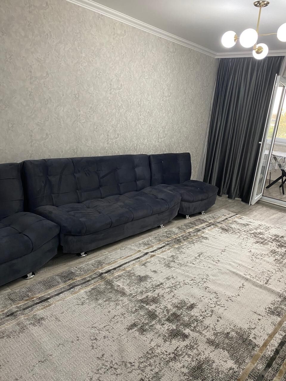 A spacious living area is featured, with a dark-colored sectional sofa positioned along a textured wall. A large area rug covers the floor, and floor-to-ceiling curtains frame a balcony door, allowing natural light to brighten the room.