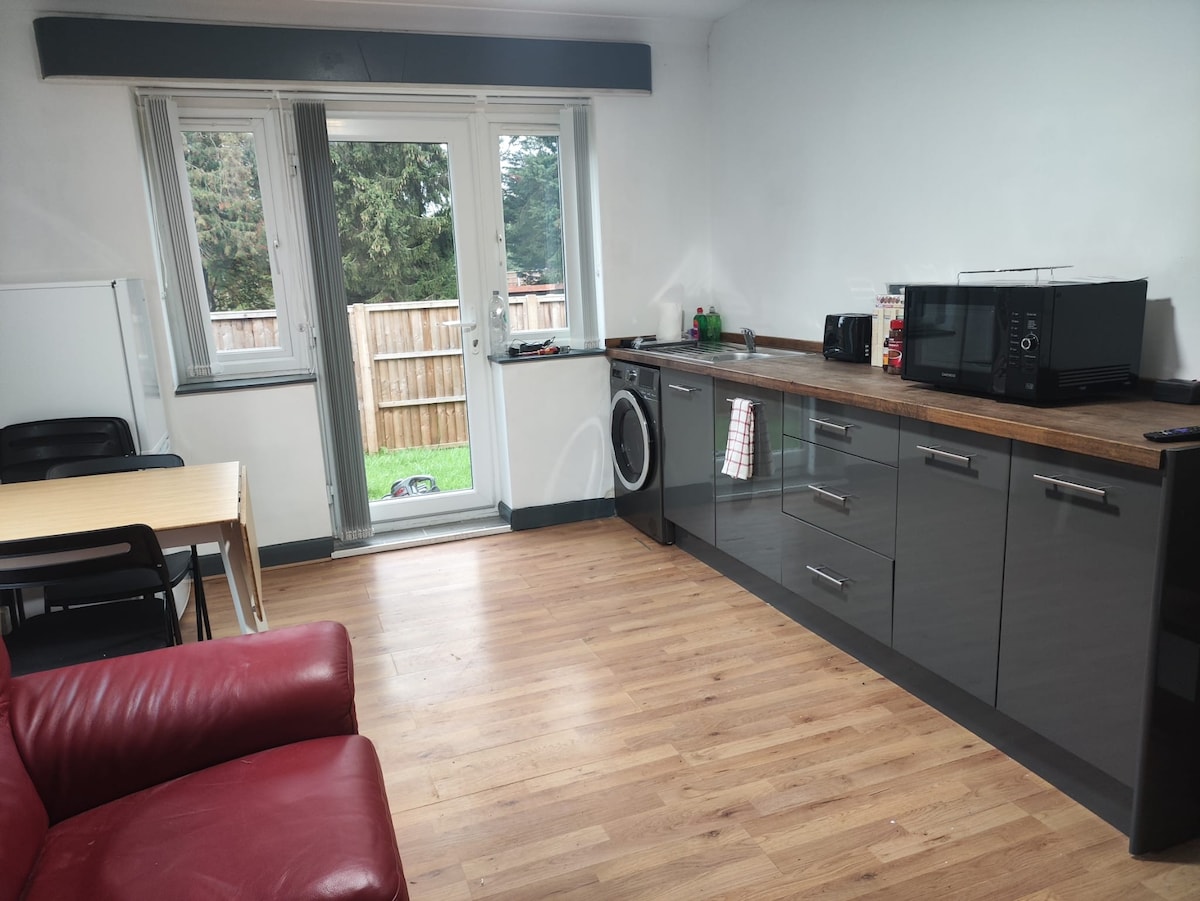 A well-equipped kitchenette is presented with sleek cabinetry and wooden countertops, featuring a microwave, kettle, and coffee machine. A dining table and four chairs are positioned nearby. Natural light enters through the glass door, which leads to the enclosed garden.