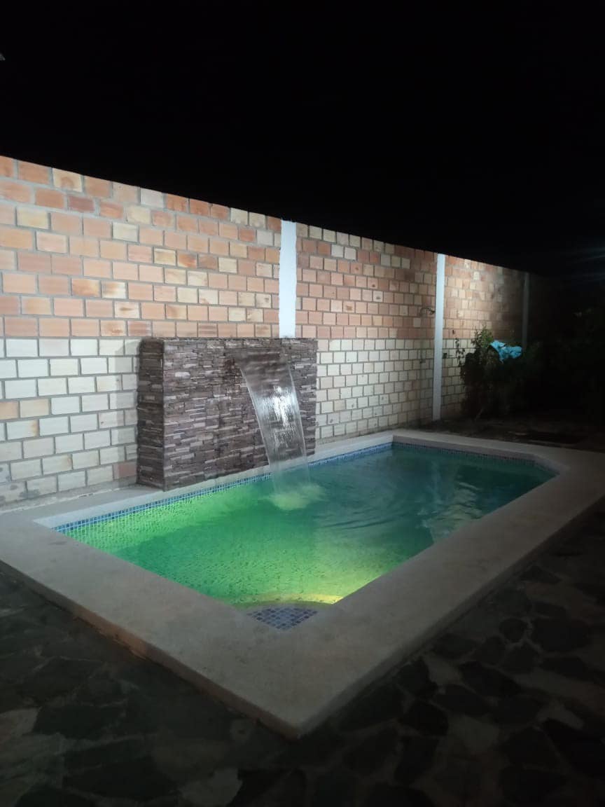A small outdoor pool is set against a textured stone wall, featuring a waterfall that adds a serene touch. Soft green lights illuminate the water, creating a calming atmosphere. Surrounding plants offer a natural feel in this private space.