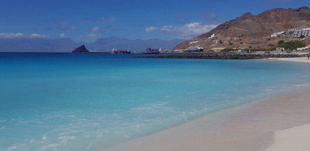 A serene beach scene is depicted, featuring soft white sand meeting clear blue waters. Gentle waves lap at the shore, with distant mountains providing a natural backdrop. The sky is mostly clear, with a few clouds adding texture, and boats are visible along the coastline.