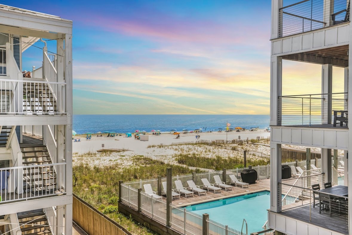 A view reveals the sandy beach along the Gulf of Mexico, dotted with colorful umbrellas and lounge chairs. The scene includes a shimmering swimming pool and the bright sky transitioning into hues of pink and orange at sunset.