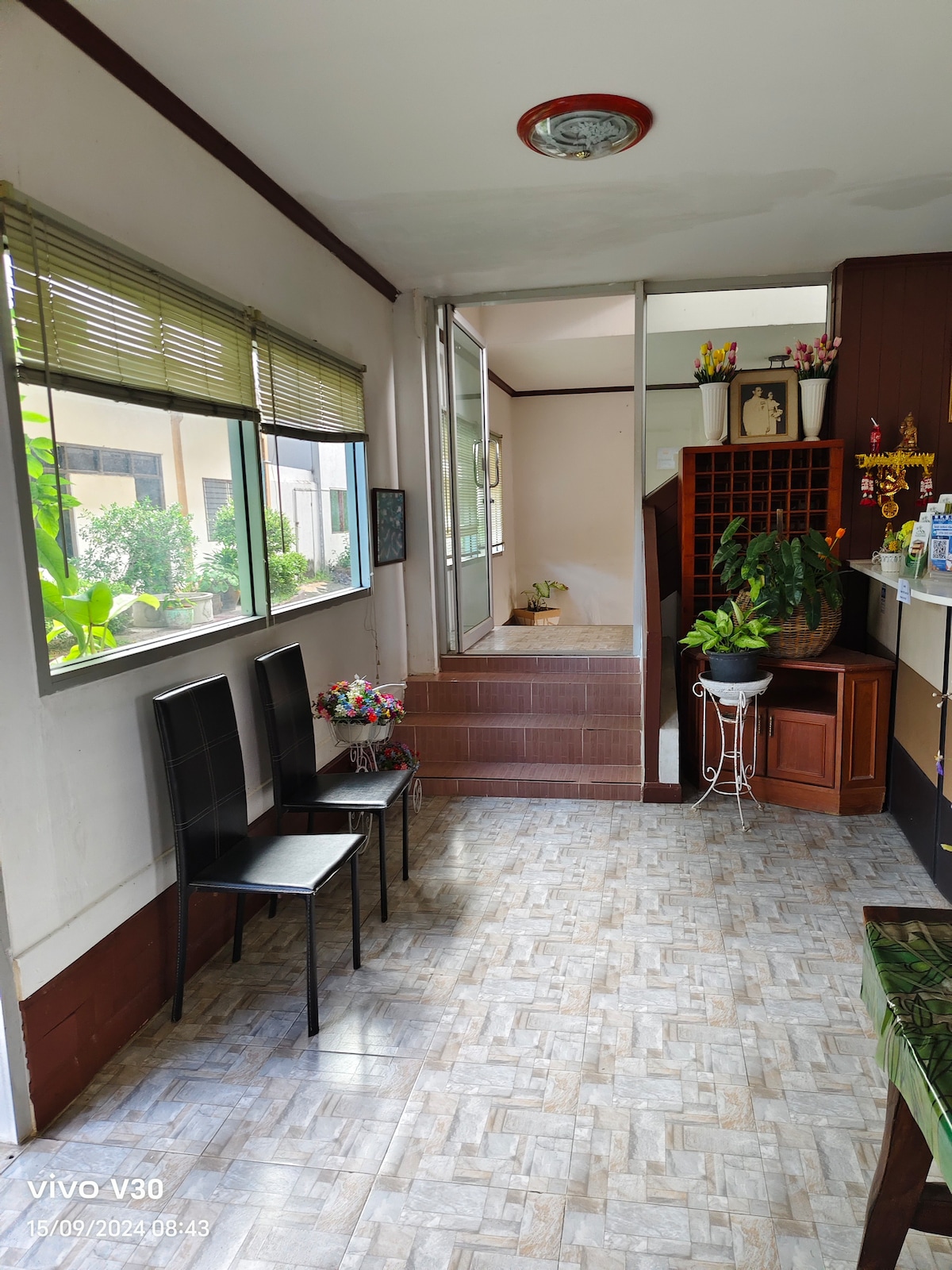 A welcoming entryway features two black chairs positioned beside a small decorative flower arrangement. Natural light enters through large windows adorned with blinds, illuminating the tiled floor and the surrounding greenery visible through the glass.