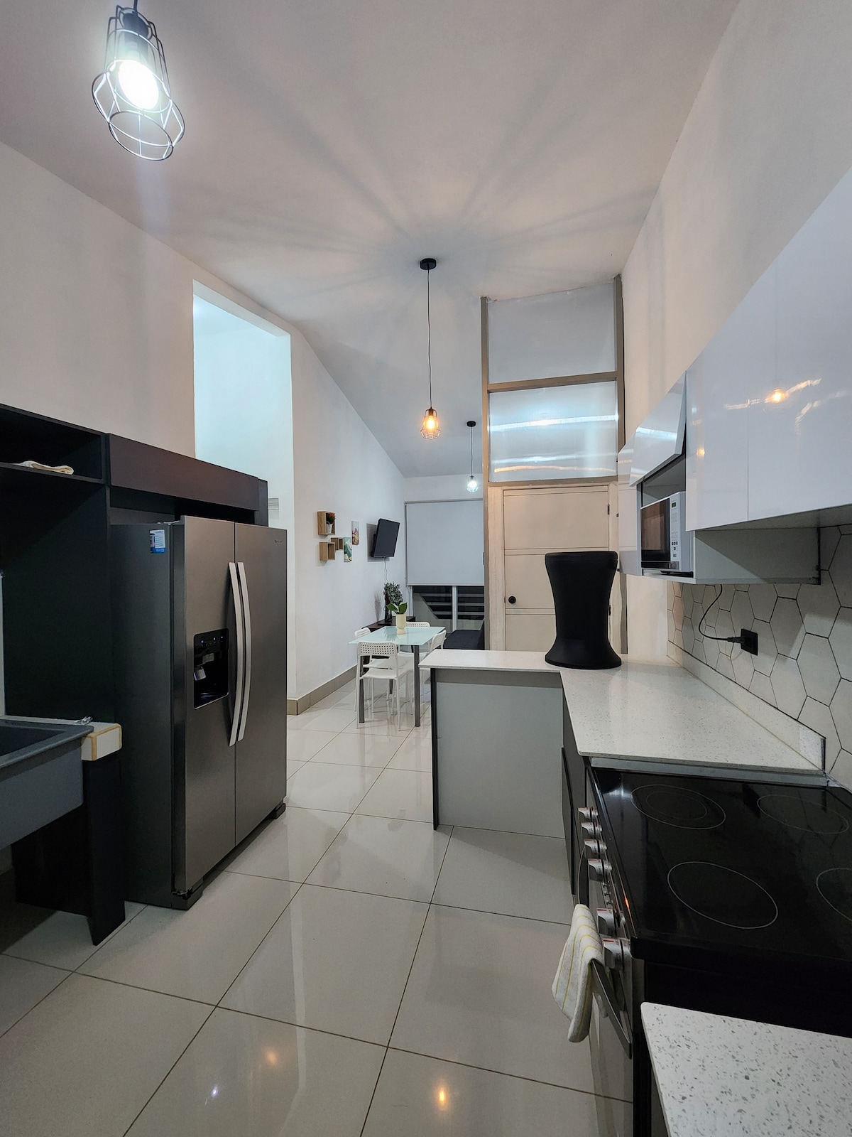 The kitchen is equipped with modern appliances, including a refrigerator and microwave. A sleek island with a cooktop is visible, complemented by a geometric tile backsplash. Ample counter space and overhead lighting create a well-lit cooking area. The open layout extends towards a living space in the background.