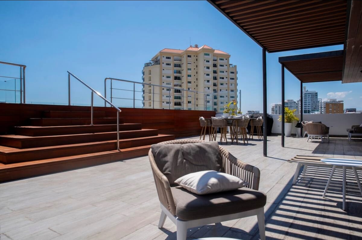 An outdoor terrace area features a wooden deck with seating arrangements, including chairs and a small table. Steps lead up to a higher platform, while the city skyline is visible in the background under clear blue skies.