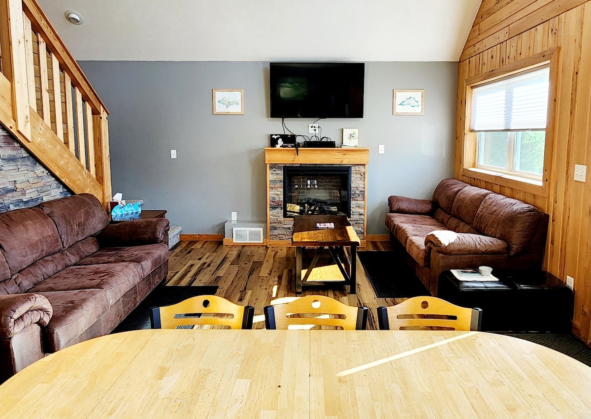 A cozy living area showcases two brown sofas positioned around a central coffee table, with a stone fireplace in the background. A flat-screen TV is mounted on the wall above the fireplace, and large windows fill the space with natural light.