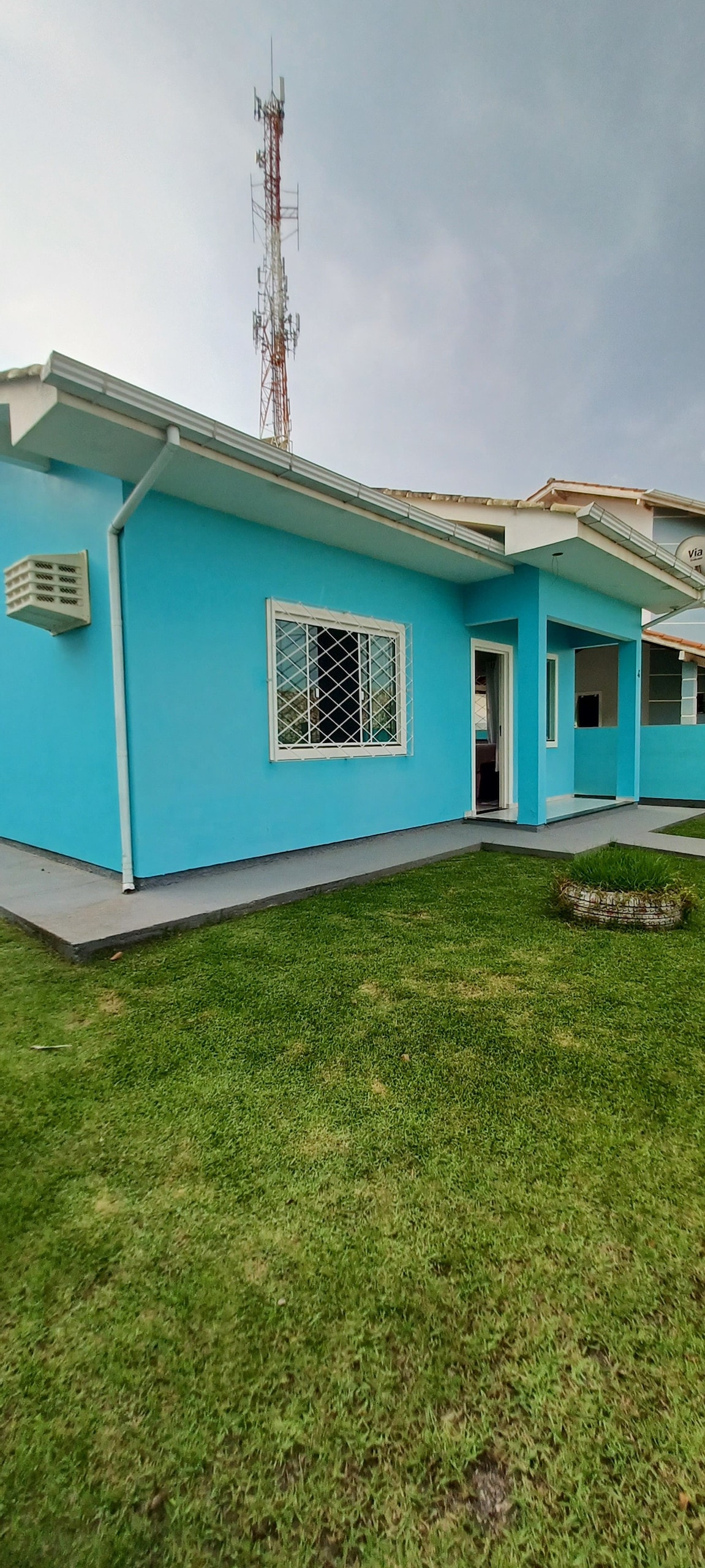 A light blue exterior of a single-story house is shown, featuring a neatly maintained green lawn. A small flower arrangement is visible on the ground, and a window with a decorative grille provides natural light to the interior.