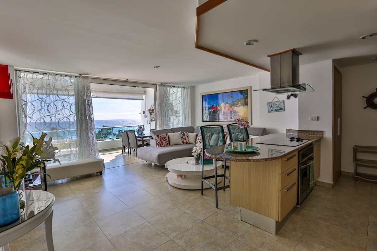 An open-plan living area is highlighted by a spacious sofa facing large windows that showcase ocean views. A modern kitchen island displays a colorful arrangement of dishes and floral decor, while natural light fills the room through sheer curtains.