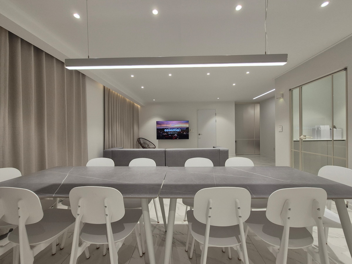 A dining area is furnished with a large table surrounded by multiple chairs. Soft lighting illuminates the space, where a cozy sofa is positioned against a wall. A flat-screen TV is mounted opposite the seating area, complemented by elegant drapes on the windows.