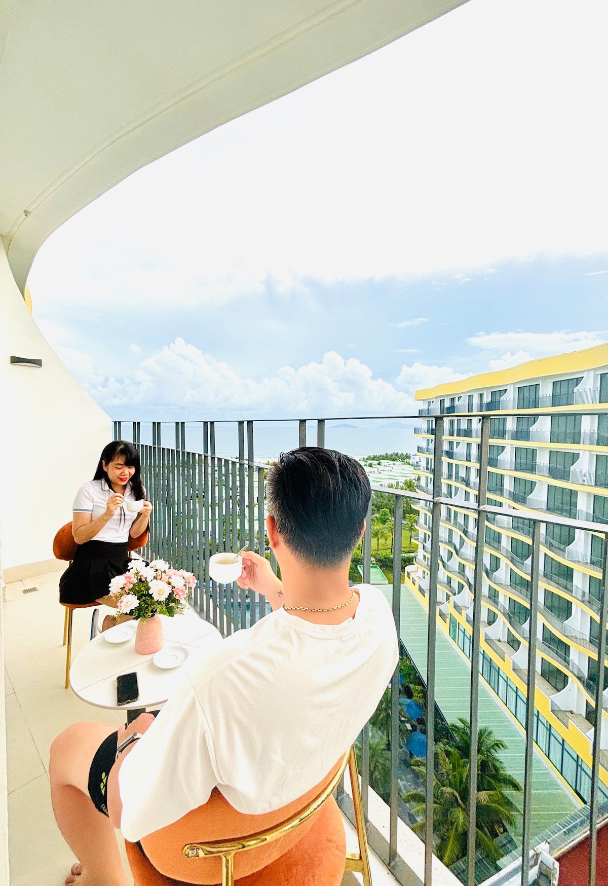 A spacious balcony is featured, overlooking the ocean and the surrounding resort. Two guests can be seen enjoying beverages at a small table adorned with a flower vase. The area is bathed in natural light, with a railing providing a clear view of the landscape.