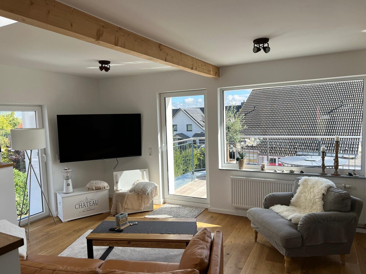 A bright living area features large windows that invite natural light and offer views of the surroundings. A comfortable gray armchair sits beside a wooden coffee table, while a flat-screen television is mounted on the wall. Light wooden flooring complements the muted color palette.