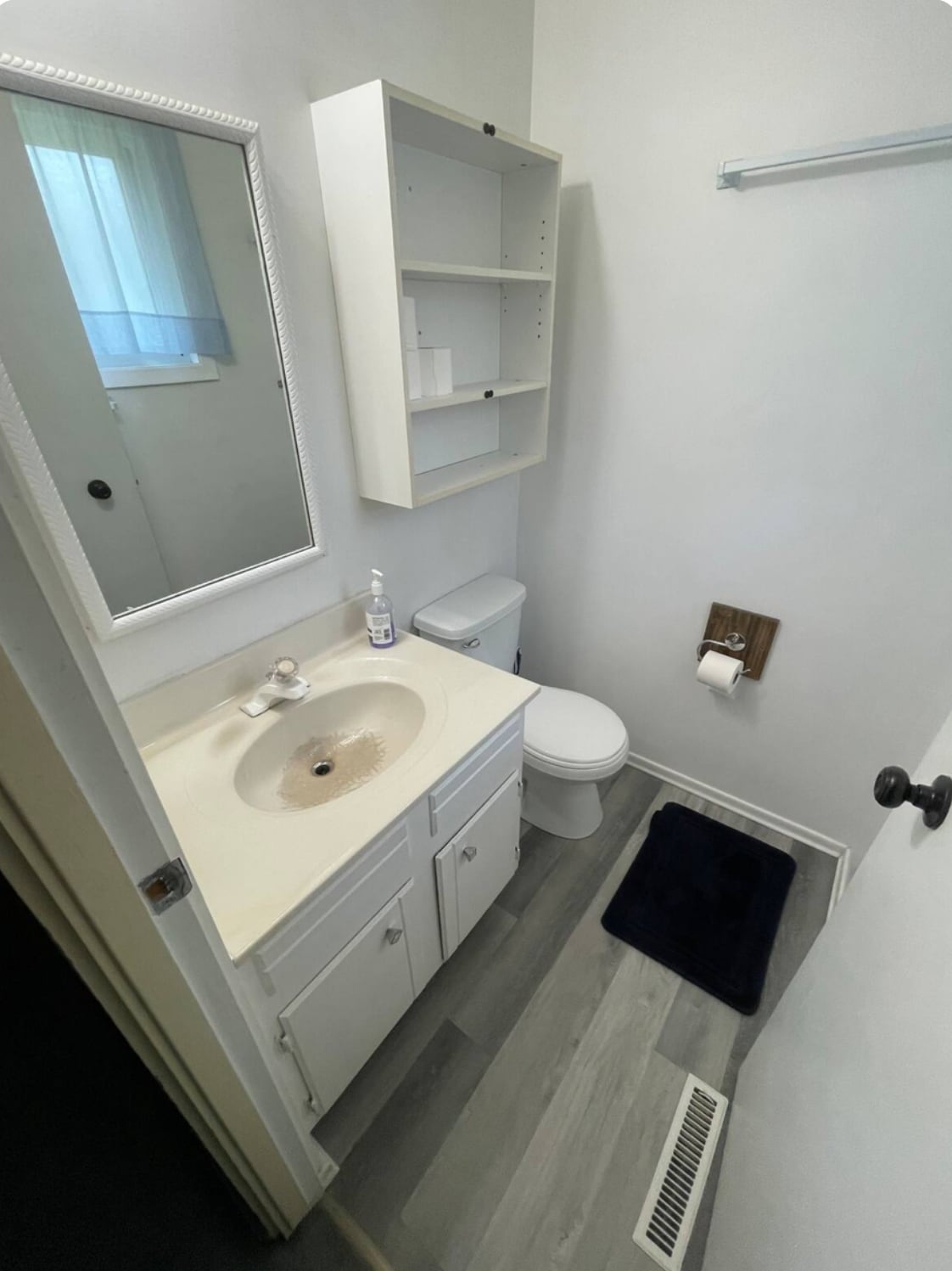 The bathroom features a modern sink with a light countertop, complemented by a wall-mounted mirror. Above the sink, simple shelving provides storage. A toilet is positioned beside the sink, and a dark mat is laid on the floor near it.