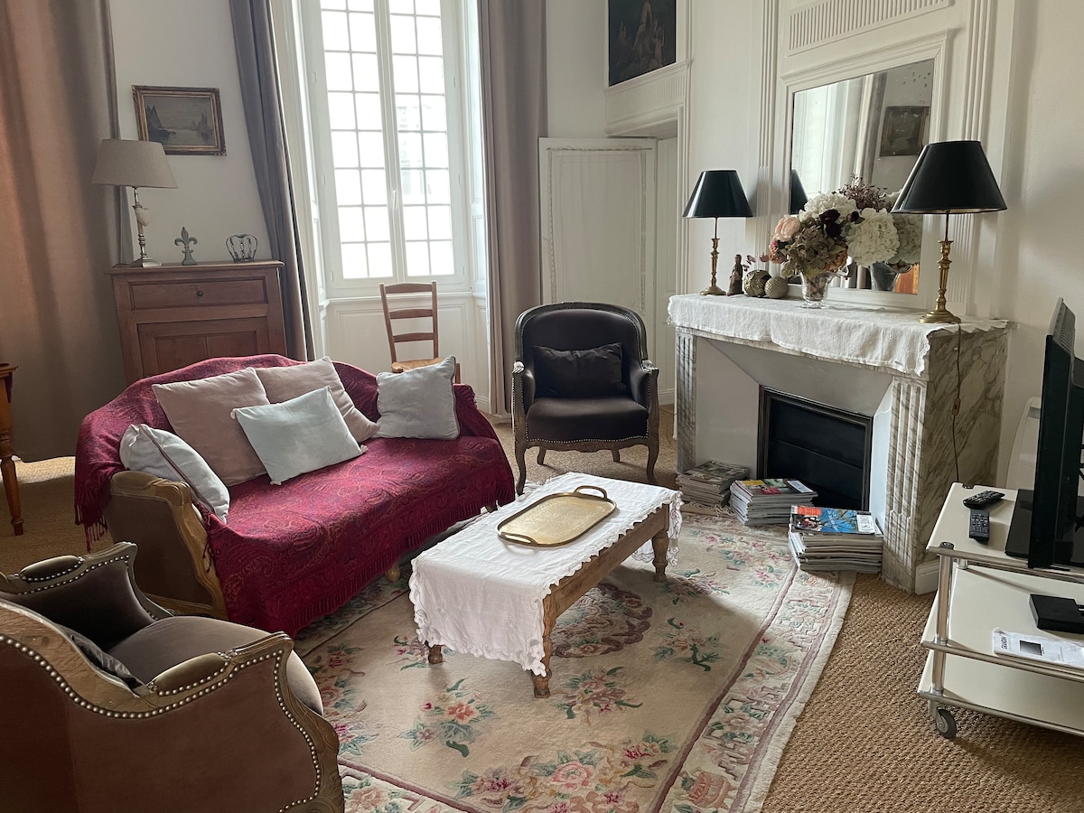 A comfortable living area features a red sofa adorned with soft cushions, complemented by a pair of elegant armchairs. A decorative coffee table rests on a patterned rug, while large windows allow natural light to illuminate the space. A marble fireplace adds a touch of charm.