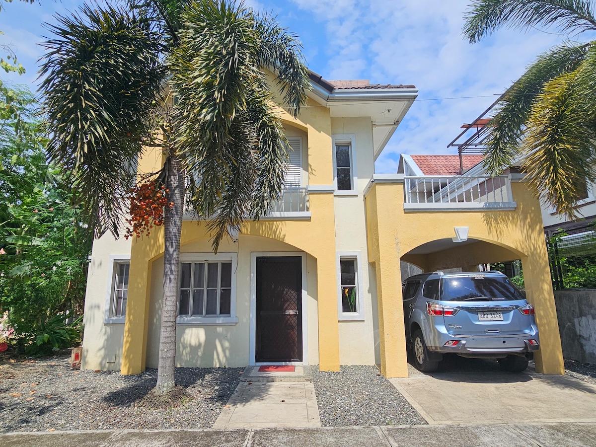 A two-story house is presented with a bright yellow exterior and a covered carport. Lush palm trees frame the entrance, while a gravel pathway leads to the front door, adding texture to the quiet environment. Windows enhance natural light, contributing to the welcoming façade.