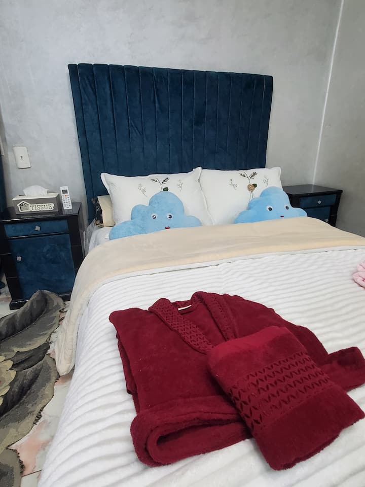 Découvrez le confort et le luxe dans cette chambre élégante. Une tête de lit en velours bleu, une literie élégante avec des oreillers nuageux et un peignoir rouge confortable créent une ambiance chaleureuse et accueillante. Chaque détail est conçu pour se détendre, pour un séjour reposant et accueillant.