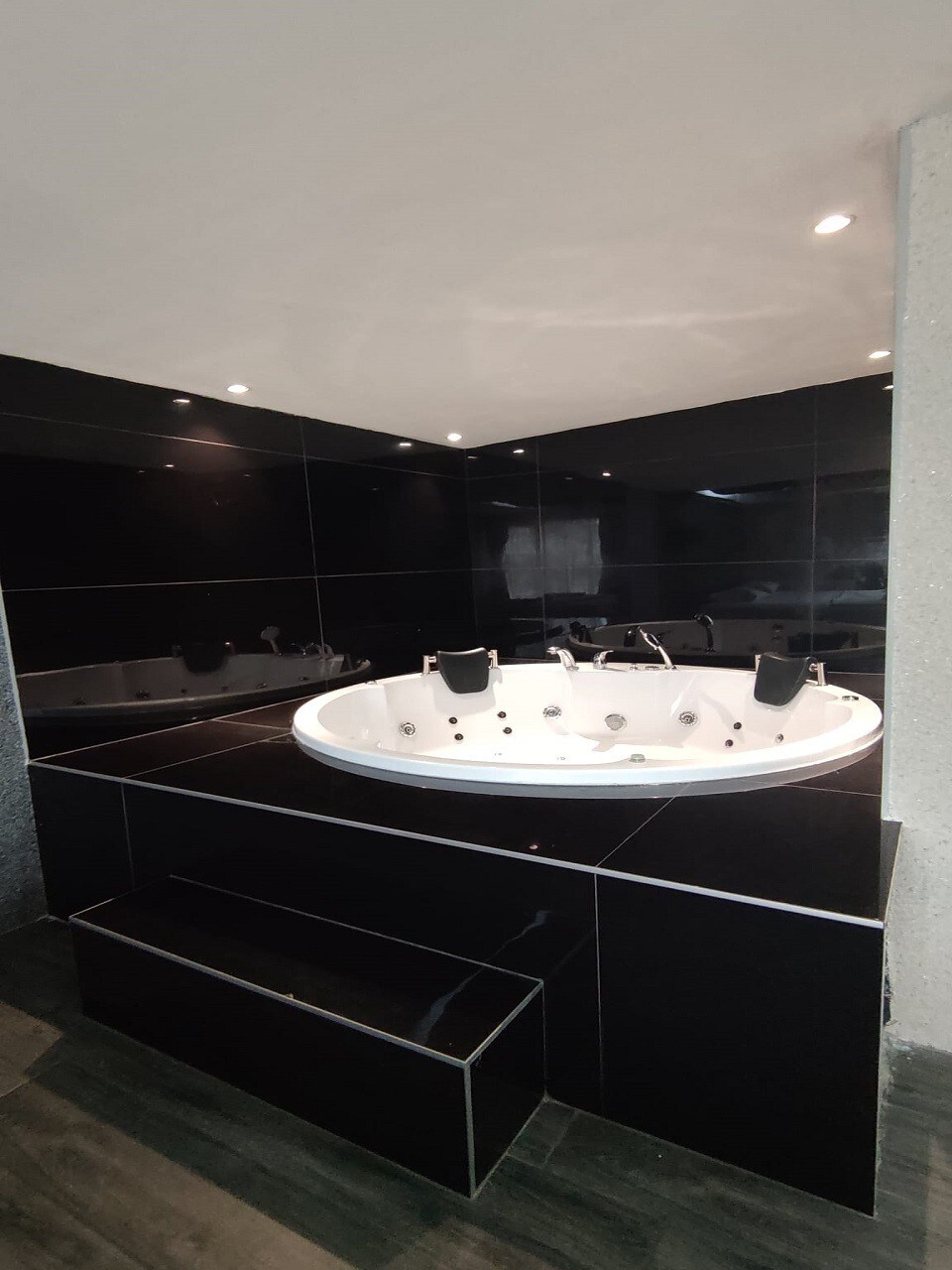 A modern jacuzzi tub is set against sleek black tile walls, illuminated by recessed lighting. The tub is accentuated by white trim and is complemented by a dark, textured floor. Steps leading up to the tub are visible, enhancing accessibility.