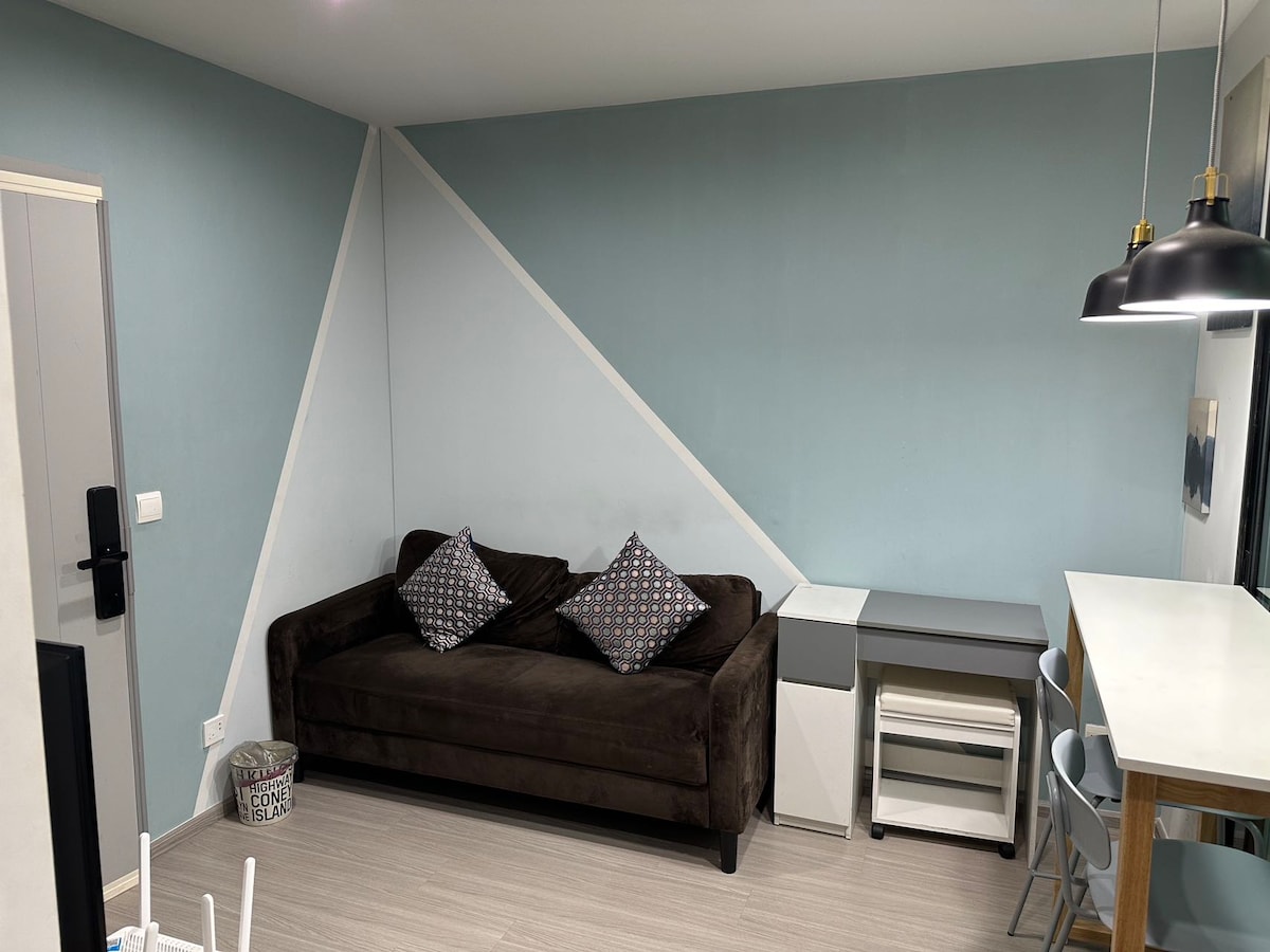 A compact living area features a dark brown couch adorned with patterned pillows. A light blue wall with a geometric design complements the space. A simple white table with chairs is positioned nearby, accompanied by a small desk and storage unit.
