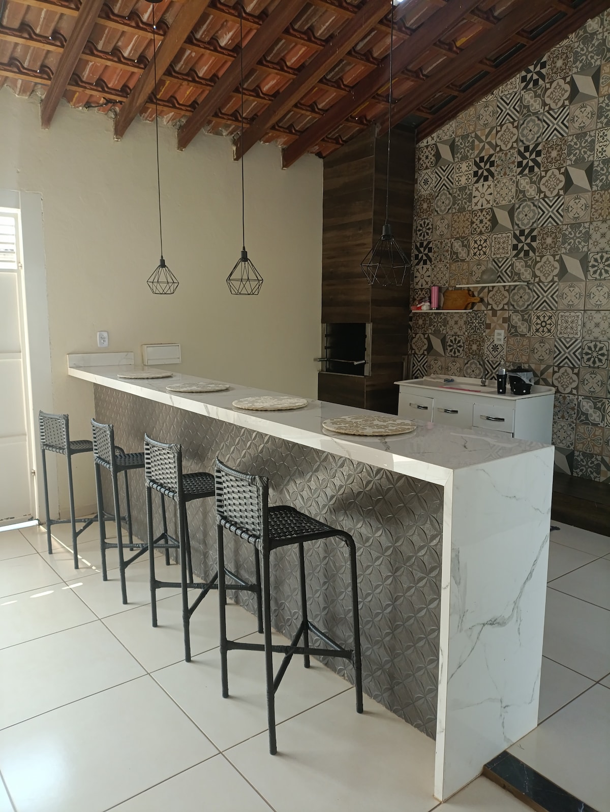 A modern kitchen features a sleek marble island with four high stools. Elegant pendant lights hang above the island, and unique patterned tiles decorate the wall behind the kitchen. A light-filled space is created by large windows that provide easy access to the outdoors.