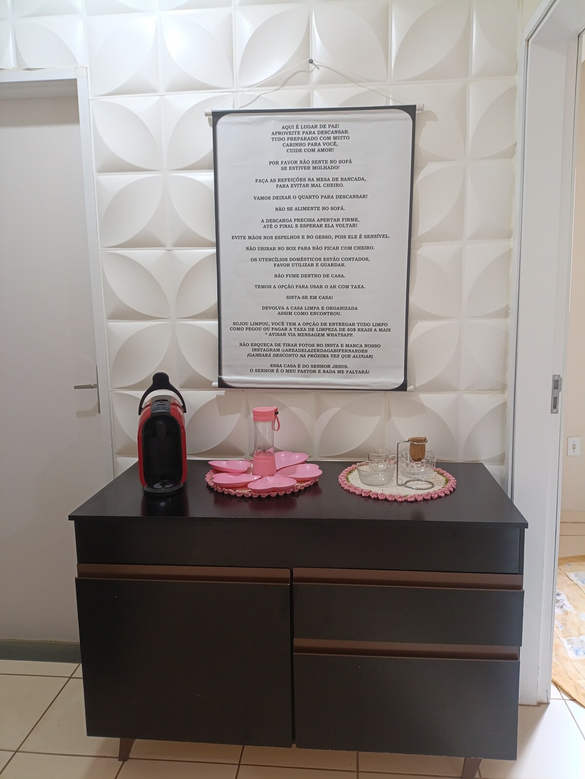 A sideboard with a dark finish is positioned against a textured wall. A decorative tray holds glasses and a pink coffee set. Above, a framed poster features text displayed on an artistic background, contributing to the area’s relaxed ambiance.