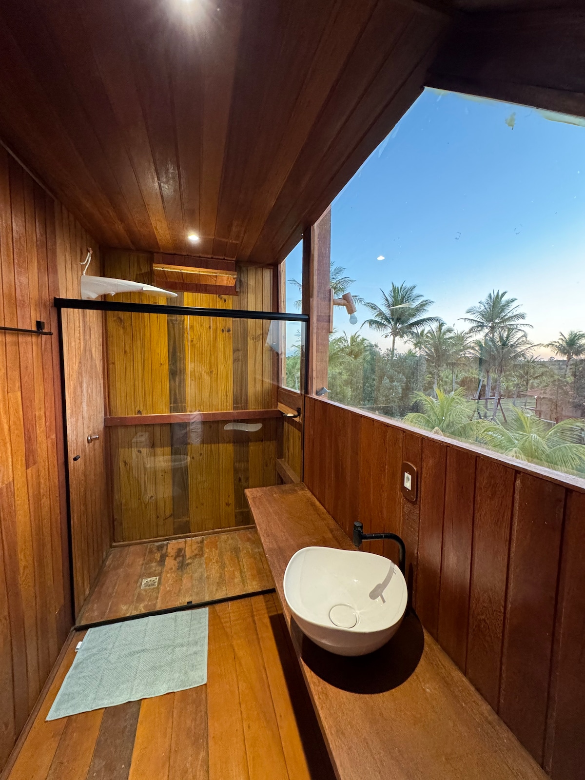 A wooden bathroom features warm tones with large windows revealing views of lush greenery. The spacious shower area is enclosed with a glass partition, and a minimalist sink is positioned on a wooden counter. Soft lighting adds to the tranquil ambiance.