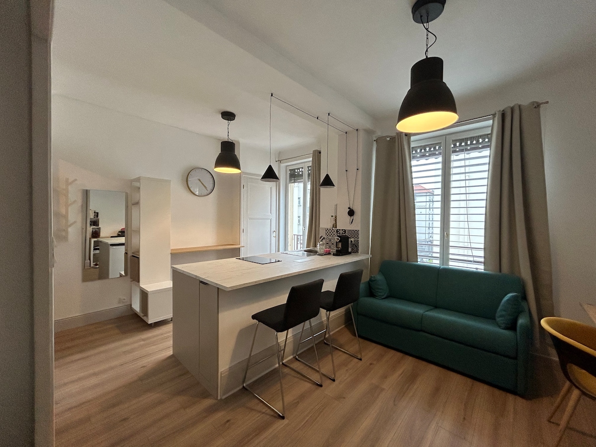 A bright and functional studio space features a spacious living area with a green sofa and three metal bar stools positioned at a modern kitchen island. Two pendant lights hang above, and large windows allow natural light to fill the room, complemented by light-colored walls and wood flooring.