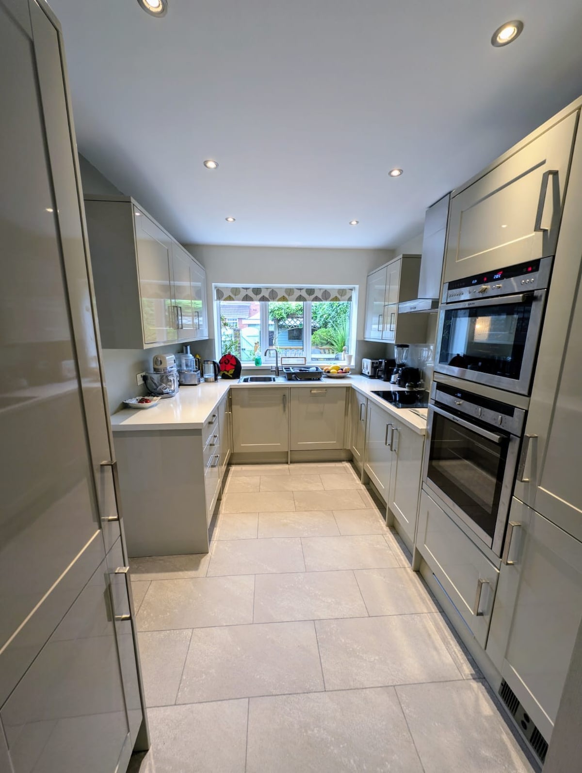 The kitchen features sleek cabinets with a modern light finish and a compact layout. High-quality appliances, including an oven and microwave, are built into the cabinetry. A window allows natural light and offers a view of the greenery outside.