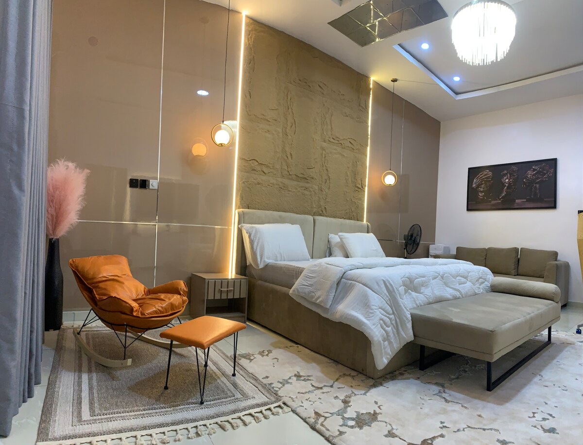 A spacious sleeping area is highlighted by a large, inviting bed flanked by softly glowing pendant lights. A stylish orange lounge chair and a small side table are positioned nearby, while an area rug adds texture to the floor. Decorative wall features enhance the overall design.