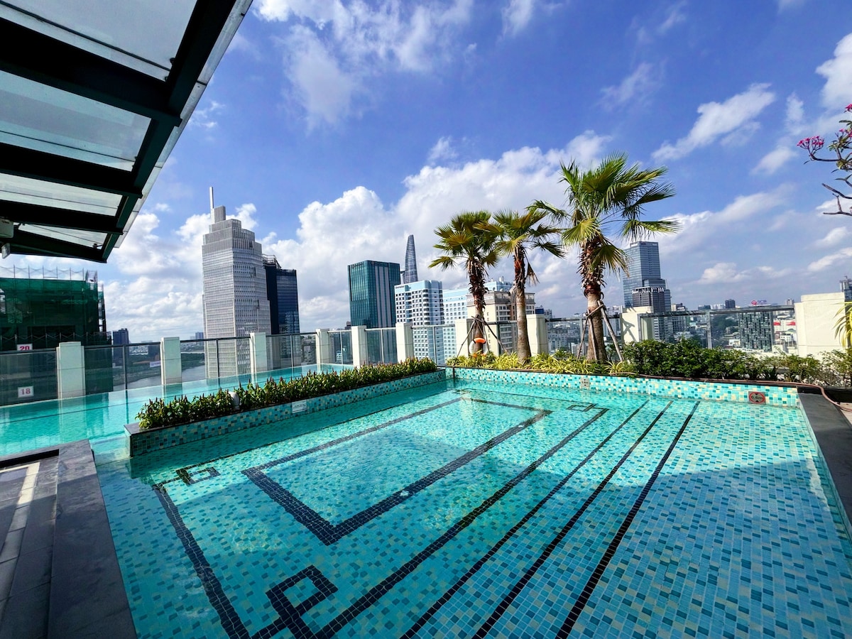 An expansive rooftop infinity pool is surrounded by vibrant greenery and palm trees. The pool features a colorful mosaic design and offers panoramic city views against a backdrop of clear blue skies and scattered clouds.