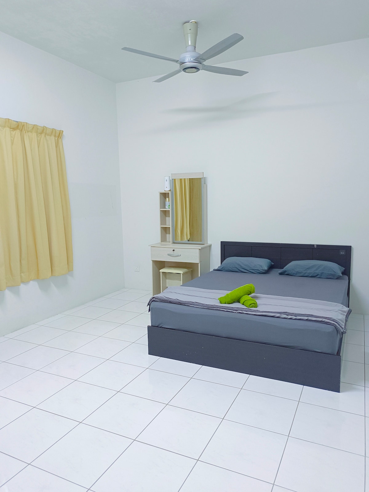 A simple bedroom is presented featuring a dark wooden bed with gray bedding. Yellow curtains frame a window, allowing natural light to fill the room. A light-colored dresser with a mirror and a towel neatly rolled is positioned on the bed, enhancing the space's functionality.